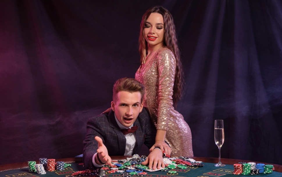 Guy is playing poker at casino sitting at table with stacks of chips, money, cards, champagne on it. Celebrating win with gorgeous woman. Black background, colorful backlights. Gambling. Close-up.